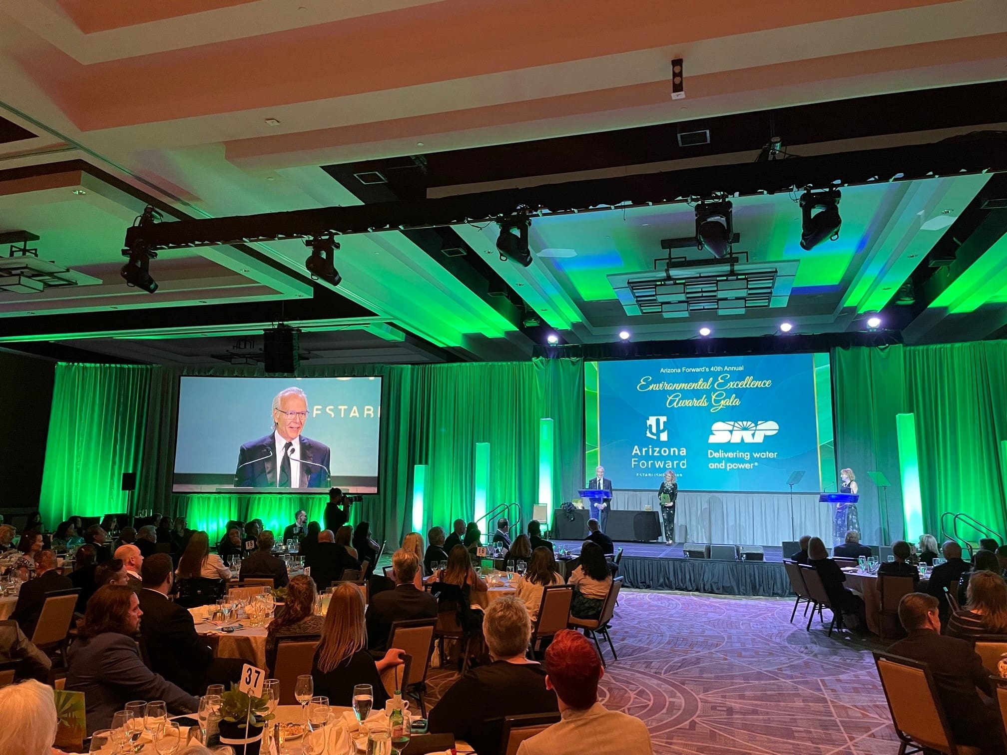 A formal event with attendees seated at tables, a speaker at a podium on stage, and presentation screens displaying "Building Better Together for a Greener Tomorrow: How Kinney Honors Earth Day" in a green-lit ballroom.