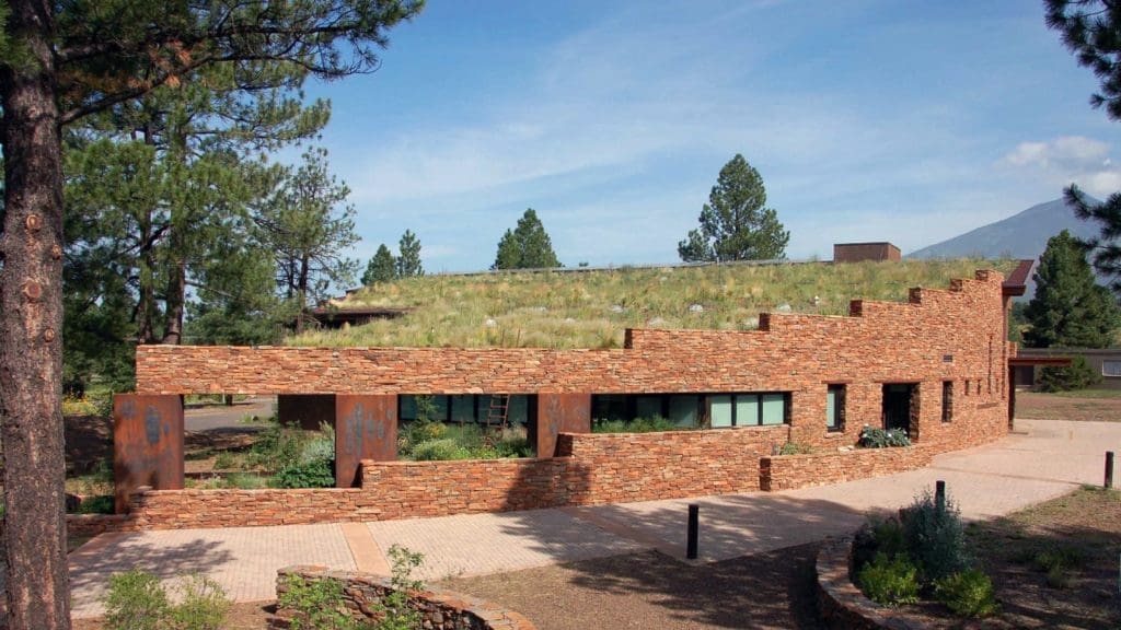 A modern, single-story building with stone walls and a green roof blends into a wooded landscape under a clear blue sky.