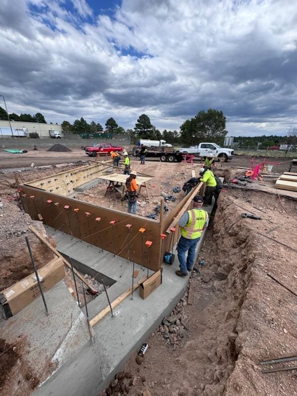 Construction workers build a concrete foundation with wooden formwork and steel rebar at an outdoor construction site under a cloudy sky.