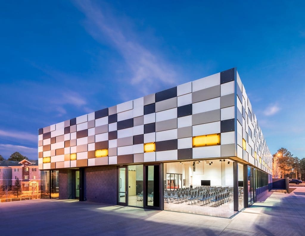 A modern building with a checkerboard-patterned facade and large windows houses the Northern Arizona University International Pavilion PV System, its illuminated interior visible through the glass, beautifully photographed at dusk.